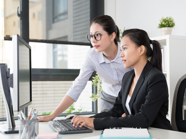 Woman seated at a desk typing on a computer while another leans over looking at the screen